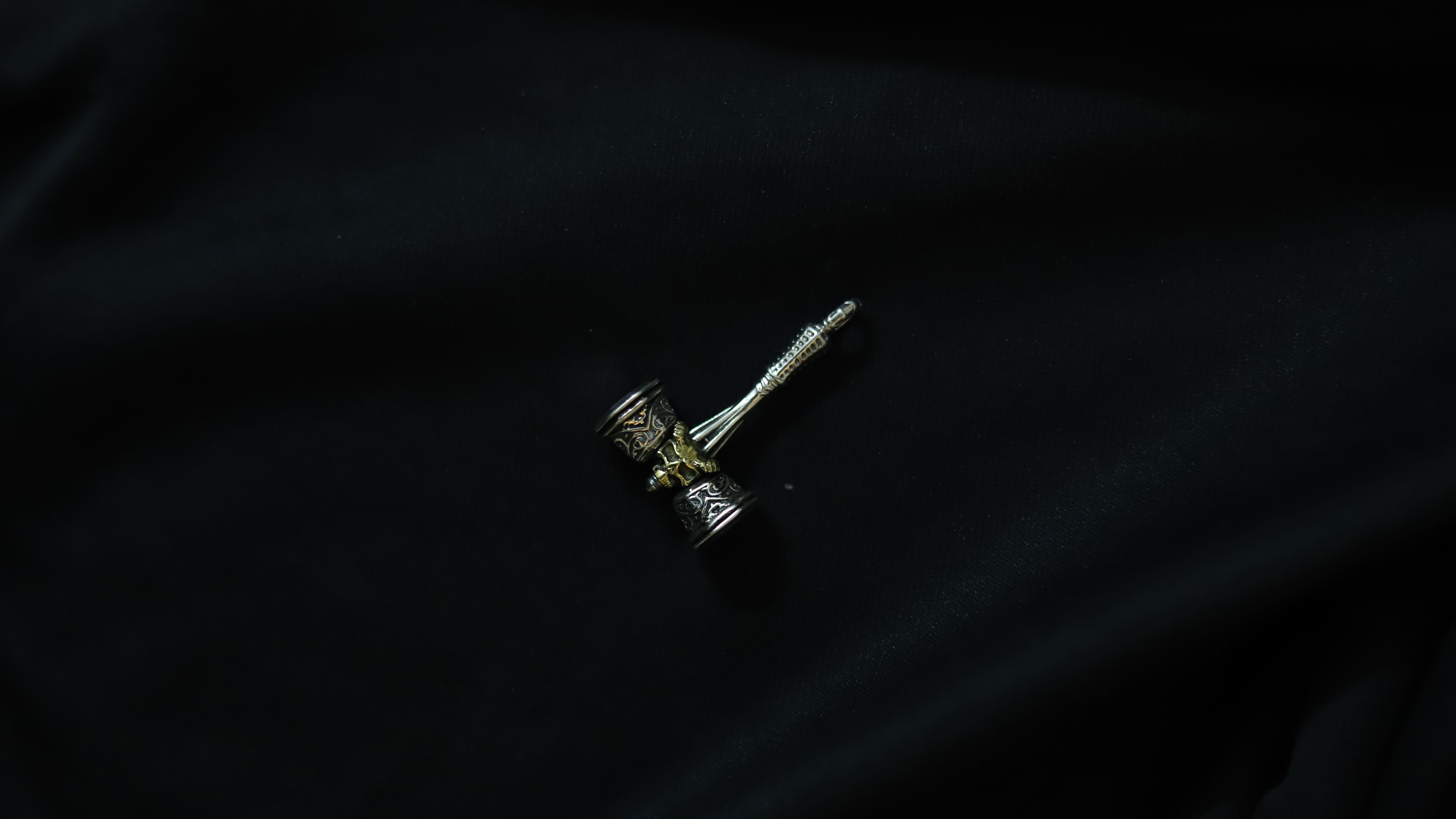 A small silver-toned metal pendant or charm with a textured cylindrical head and a long pointed stem, photographed on a dark black fabric background.